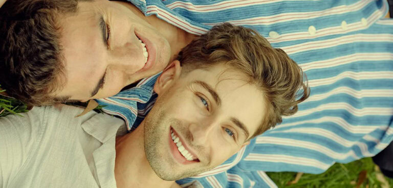 Two young men lie on the grass, both wearing collared shirts, they smile, squinting in the sunshine. One has his head on the other's chest.