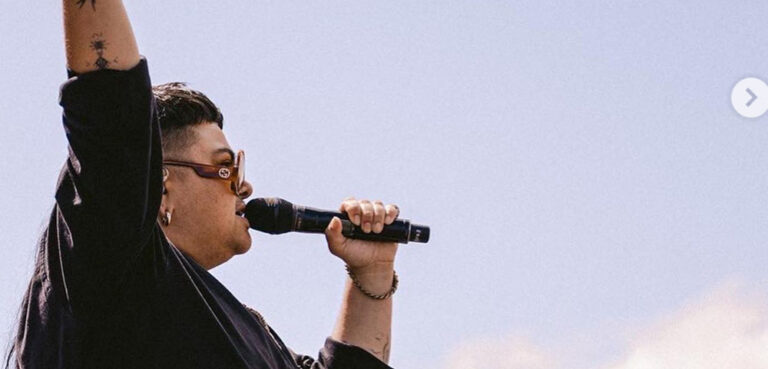 Mo'Ju singing into a microphone with their hand raised against a warm blue sky