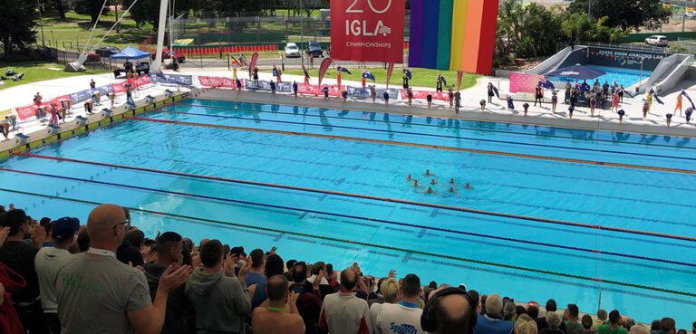 A large pool photographed from a distance, the crowd is full and a rainbow flag hangs over the water