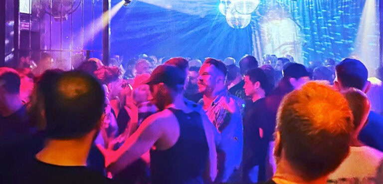 Image shows a crowd of men dancing in the blue light below a disco ball.