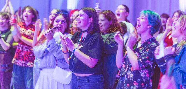 An image of a group of young LGBTQIA+ people cheering and clapping, the image has a purple filter.