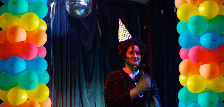 A collage image of a drag king on stage holding a microphone, overlayed with superimposed birthday hat and two rainbow chains of balloons, one on either side of the image.