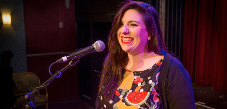 A person with long wavy brown hair wearing red lipstick smiles widely into a microphone, wearing a dress covered in fruit and a black cardigan, the light on their face is purple and orange.