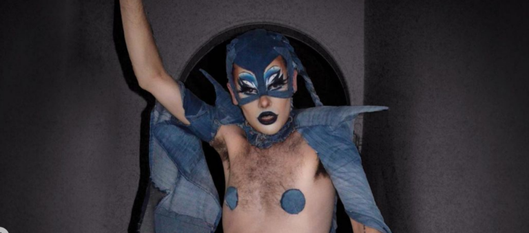 A person wearing a denim balaclava, capelet and pasties with blue lipstick and eyeshadow has their left arm raised on a dark background, staring at the camera.