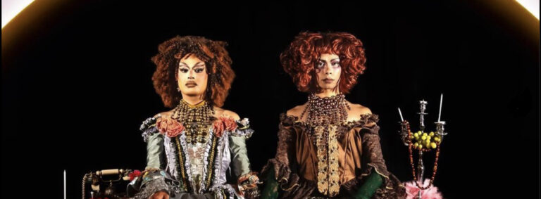 Two drag queens, Cerulean and Stone Motherless Cold, sit side-by-side wearing ballgowns and red curly hair. Cerulean on the left is wearing powder blue dress with lace and a large brass pearl collar/ruff, Stone Motherless Cold on the right is wearing a bronze/copper coloured gown with gold lace detailing and emerald green sleeves from the elbow down, and a bronze/copper pearl necklace/ruff. Their expressions are very neutral, and they are sitting between an old-style telephone and a silver candelabra adorned with green grapes and a red beaded necklace.