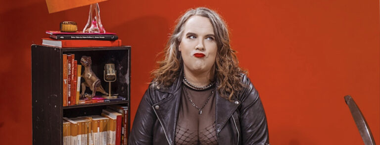 Anna, a comedian with long reddish hair and a cheeky grumpy expression, wears a black leather jacket and mesh top on a background of bright red which matches her lipstick. On her left is a bookshelf with a wonky lamp on top, a shelf with a t-rex toy and a old-school microphone, and another shelf full of orange Penguin books.