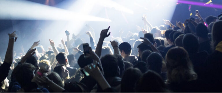A nighclub bathed in bright light, a crowd dancing with many people with their hands up in sillhouette