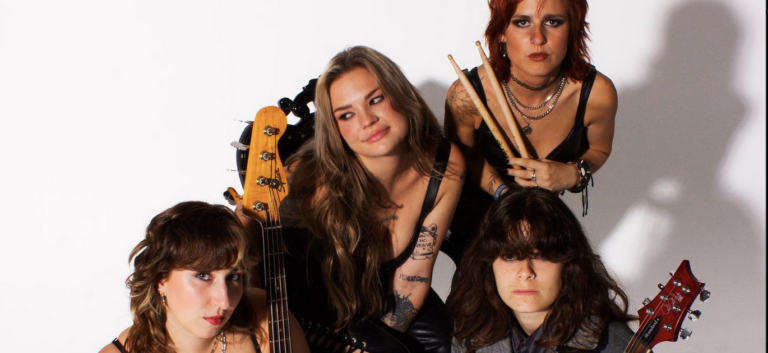 Four punk musicians on a white background, they are sitting in a group, wearing black and the two in singlets are showing tattoed arms. In the centre is the singer, looking cheekily to the left of frame, to the bottom left and right, and behind her on the top right the other musicians are looking boldly at the camera while holding a guitar, bass and drumsticks respectively.