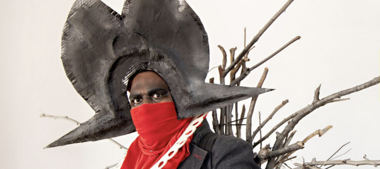 Performer Jay Wymmara wears an oversized 2d hat with a large pointed 'brim' and a red bandanna over his nose, mouth and chest, with a stylised white 2-d ruff over the top. Behind Jay are a bunch of sticks and small branches radiating out from behind Jay's shoulders.
