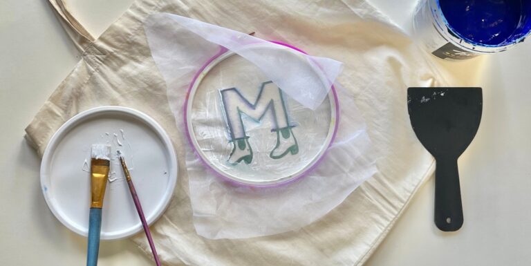 A birds eye view of a table, with a paper plate to the left, holding two brushes, a tote bag with an embroidery hoop holding a piece of paper with the letter 'M' on it, (the M is wearing little heeled cuban boots) and to the right of the image a plastic paint scraper and the edge of a paint pot with bright blue paint in the top right corner of the image.