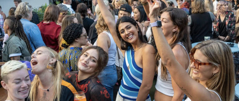 A crowd shot from a rooftop party focusing on a group of friends at the front of the crowd smiling with their hands up.