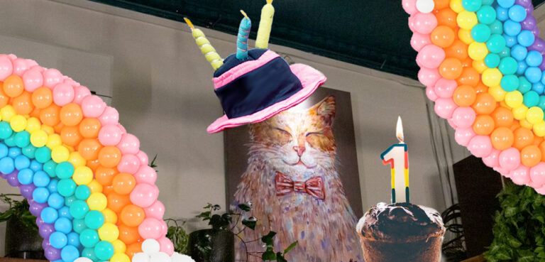 An image of a painting of a cat on a wall, it has been photoshopped to be wearing a cake-shaped birthday hat with three candles on it, there are arcs of rainbow balloons in the bottom left and top right corners, and a cupcake with a 1-shaped candle in the centre-bottom right