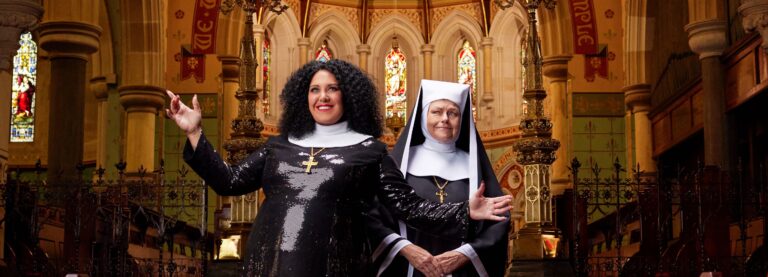 Two actresses dressed as nuns in sparkly black costumes stand in a church smiling at the camera. To the left, Casey Donovan wears her curly black hair out and has her arms outstretched, smiling, to the right Genevieve Lemon is wearing a black and white wimple and has a sterner expression.