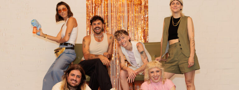 Six people stand and sit (on two green chairs or the floor) in front of a gold fringe backdrop. They are all smiling and wearing casual, playful clothing. One on the far left is holding a bubble gun.