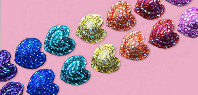 A line of pairs of heart-shaped rhinestoned nipple pasties crosses from bottom left to top right of frame, in dark purple, blue, green, yellow, orange, red and pink on a pastel pink background.