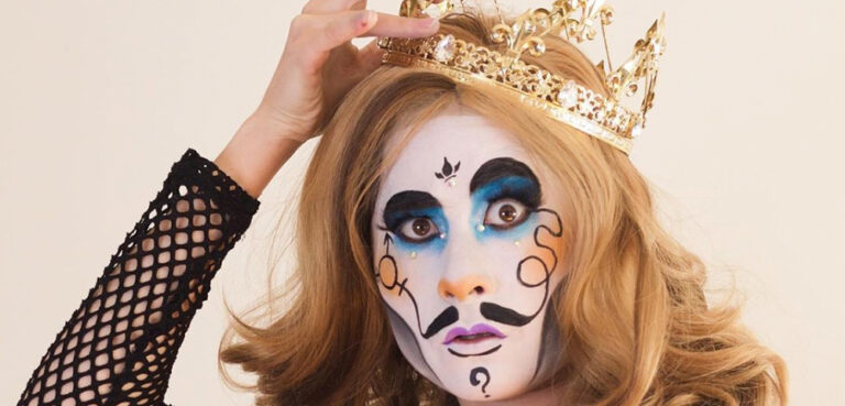 A drag king in stylized white makeup and a blonde curly wig holds a crown onto his head with one hand while looking at the camera with a bewildered expression