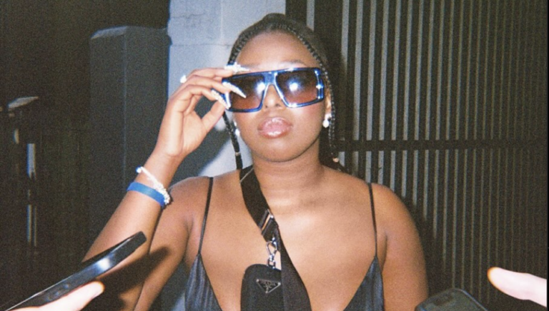 Pop musician Kye, a Zimbabwean-Australian woman, walks on the street wearing a spaghetti strap black top or dress, holds her large sunglasses on her face with her right hand, you can see two hands holding cellphones as if recording her speaking. The image looks like it is shot on film- a candid paparazzi style shot.
