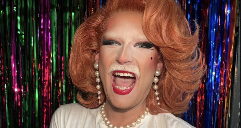 Tilly Capulet, a drag queen with a white mustache and red curly wig, wears pearl earrings and a necklace, red lipstick and a huge open-mouthed smile in front of a multicolour tinsel curtain